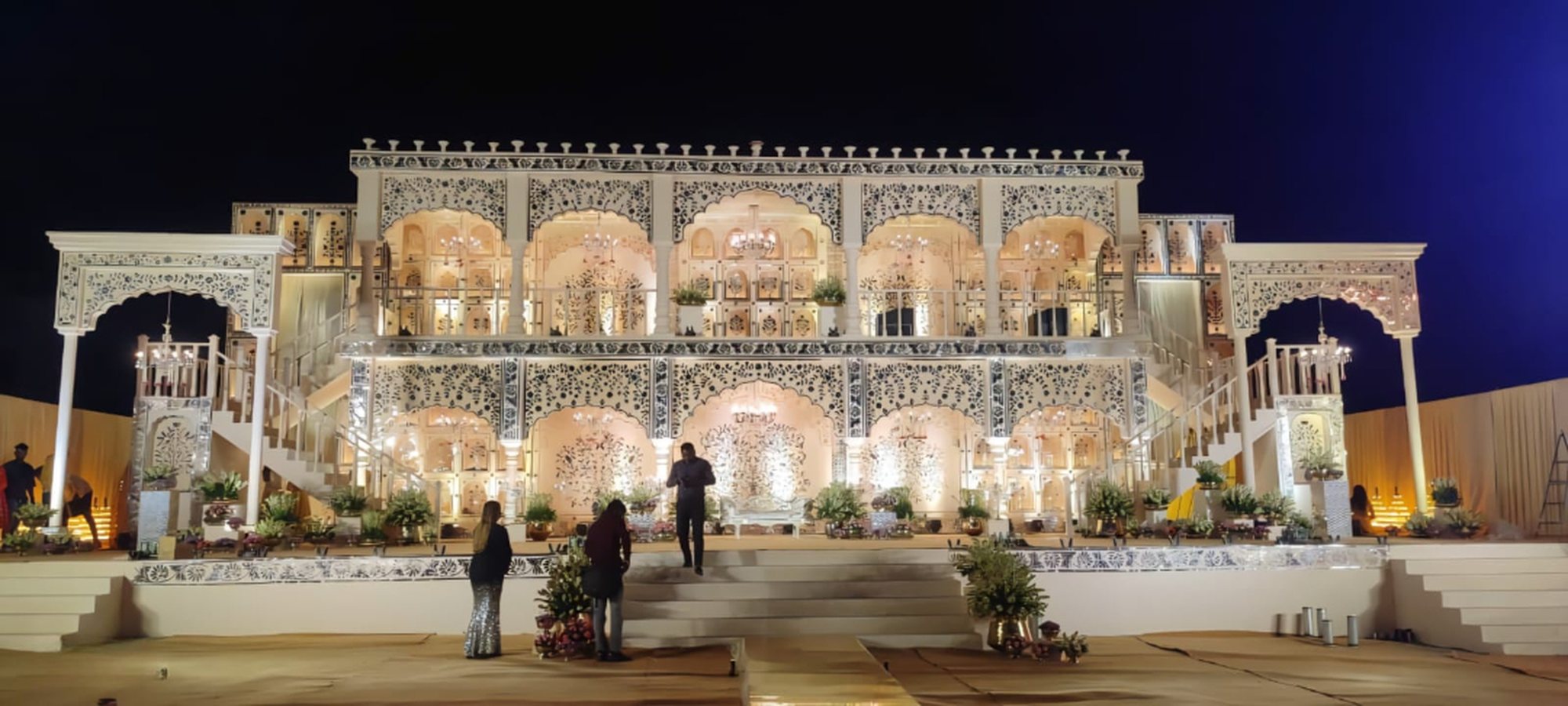 Two-storey palace stage with carved jaali arches, chandeliers and twin staircases, lit up at night